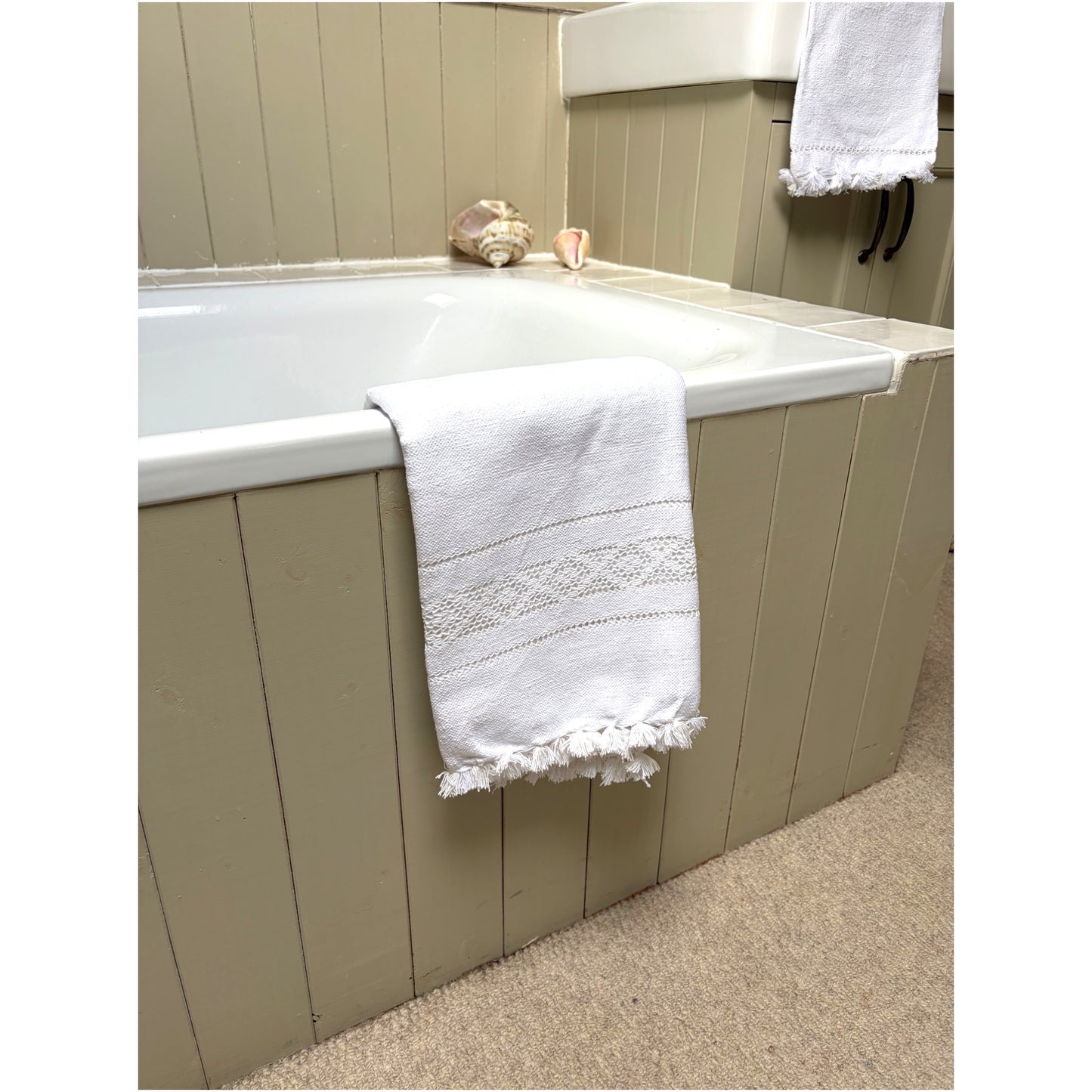 A white cotton bath towel with a decorative embroidered band and fringed edges draped over the side of a bathtub in a beige-paneled bathroom.