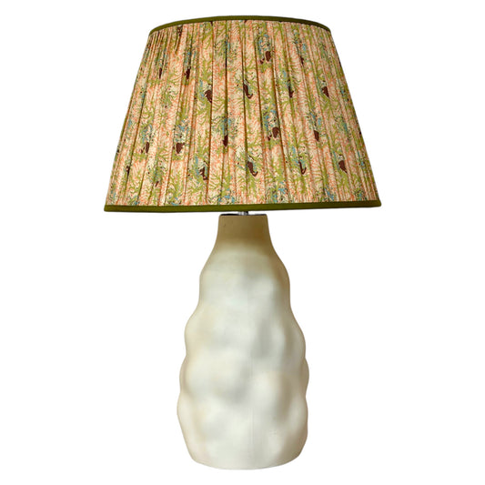 A table lamp with a textured, wavy off-white ceramic base and a gathered silk lampshade featuring a vintage coral and seaweed botanical pattern with green leaves, peach and pink flowers finished with olive green trim at the top and bottom.