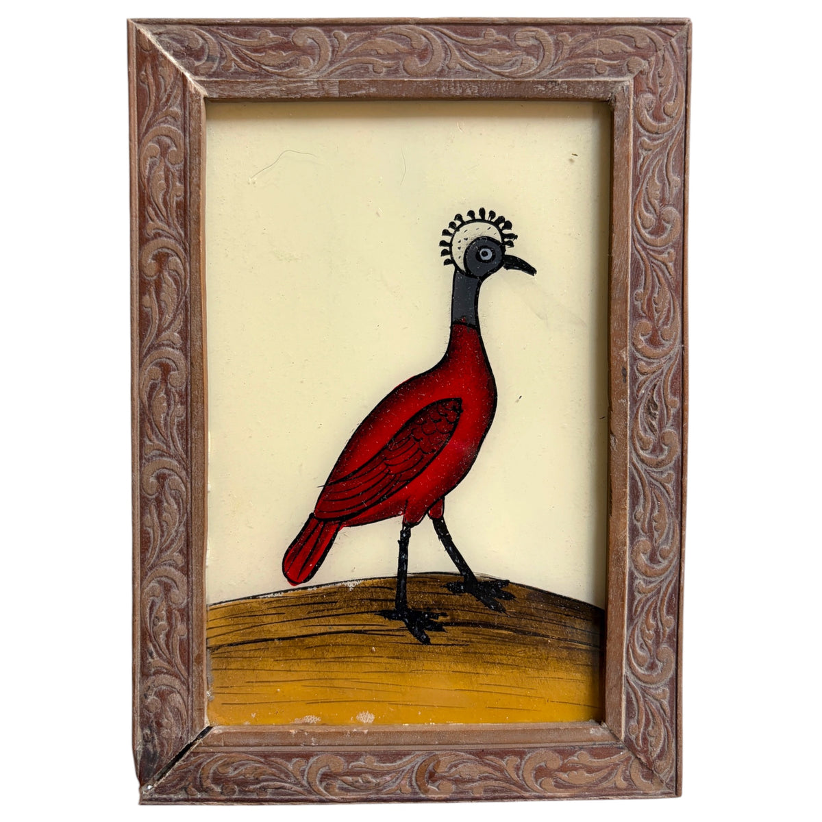A framed folk art piece featuring a stylized red bird with a black neck and crown, standing on a yellow-brown ground against a cream background, enclosed in an ornate carved brown wooden frame.