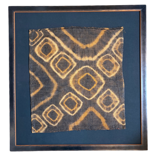 A square framed textile art piece featuring a dark charcoal background with a tan and ochre geometric diamond pattern, set in a dark wood frame with a black mat mount.