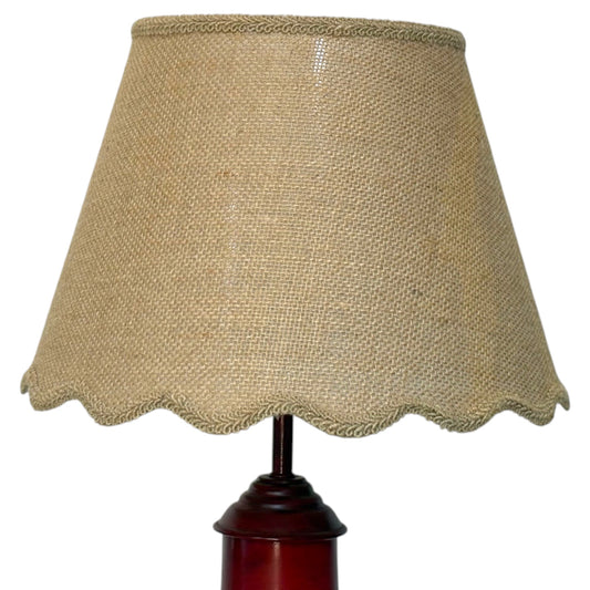Close-up of a table lamp with a natural beige hessian shade featuring a scalloped edge on a dark reddish-brown wooden base, shown against a plain white background.