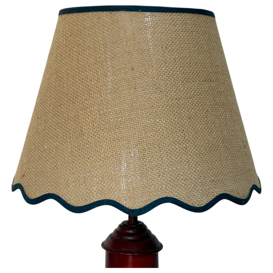 Close-up of a natural hessian lampshade with a dark blue scalloped trim, mounted on a dark reddish-brown wooden lamp base against a plain white background.