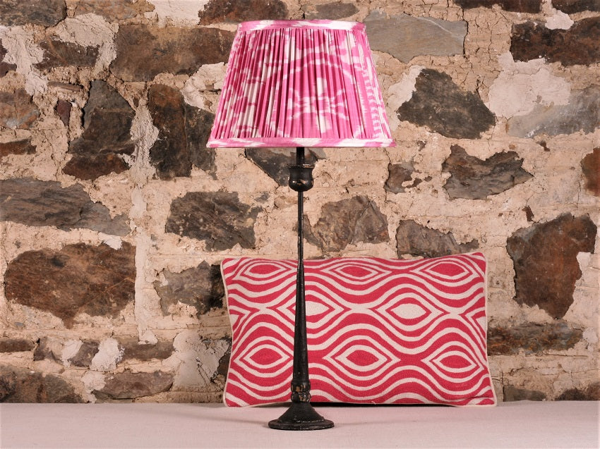 Pink Silk Ikat Lampshade shown on a candlestick lamp base in front of a stone wall and accent cushion in the background