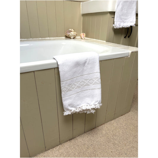 A white cotton bath towel with a decorative embroidered band and fringed edges draped over the side of a bathtub in a beige-paneled bathroom.