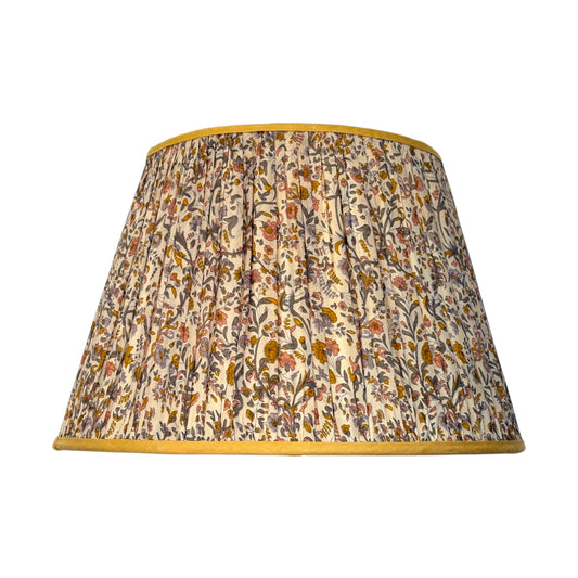 A gathered silk lampshade featuring an intricate vintage paisley floral and botanical pattern in muted tones of orange, yellow and brown on a cream background, finished with a contrasting mustard yellow trim.