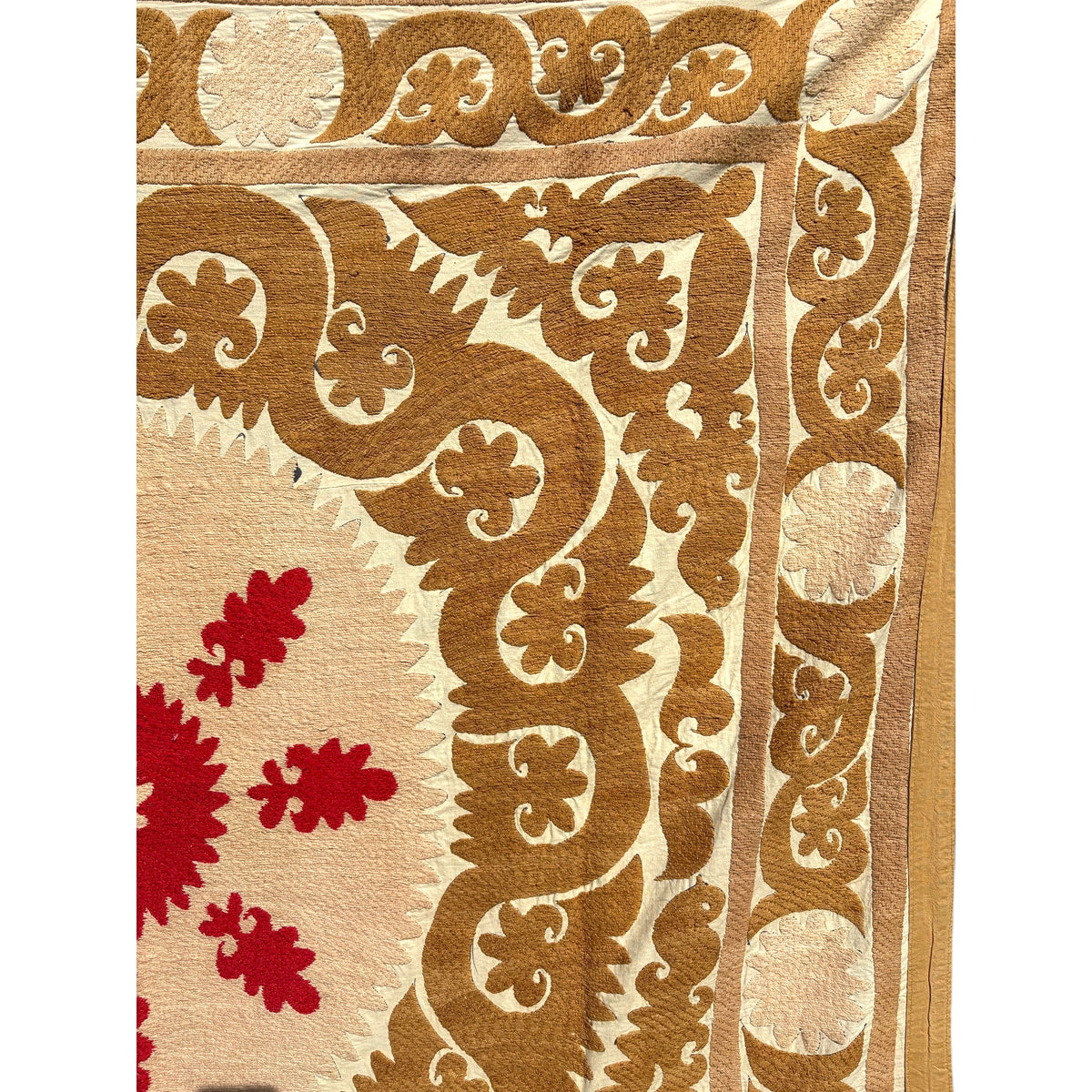 A close-up of a patterned vintage Suzani  showing a cream background with a large tan scrollwork border and red floral motifs in the lower left.