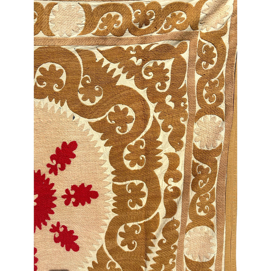 A close-up of a patterned vintage Suzani  showing a cream background with a large tan scrollwork border and red floral motifs in the lower left.