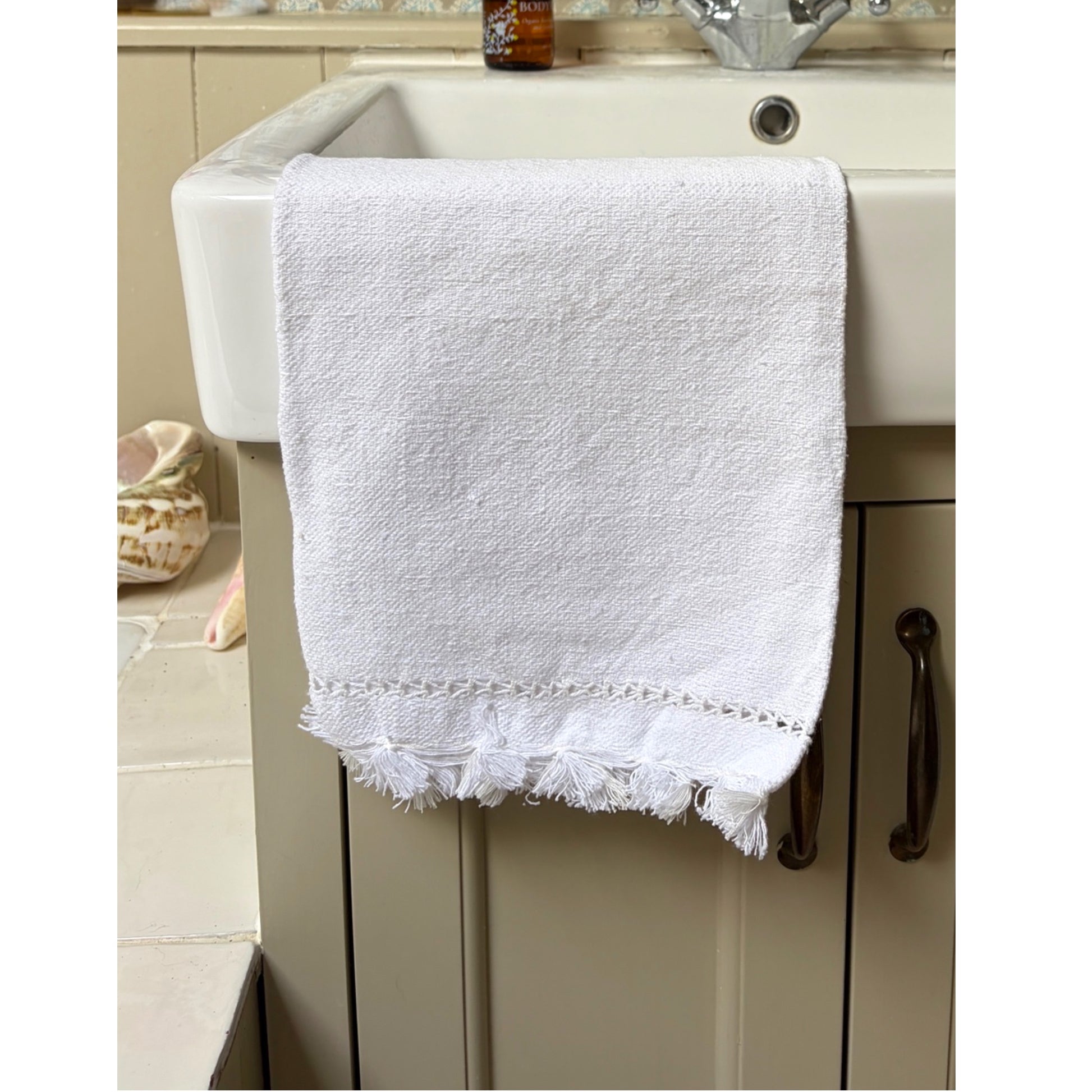 A white textured cotton hand towel with fringed tassels hanging over a white ceramic sink on a beige vanity cabinet.