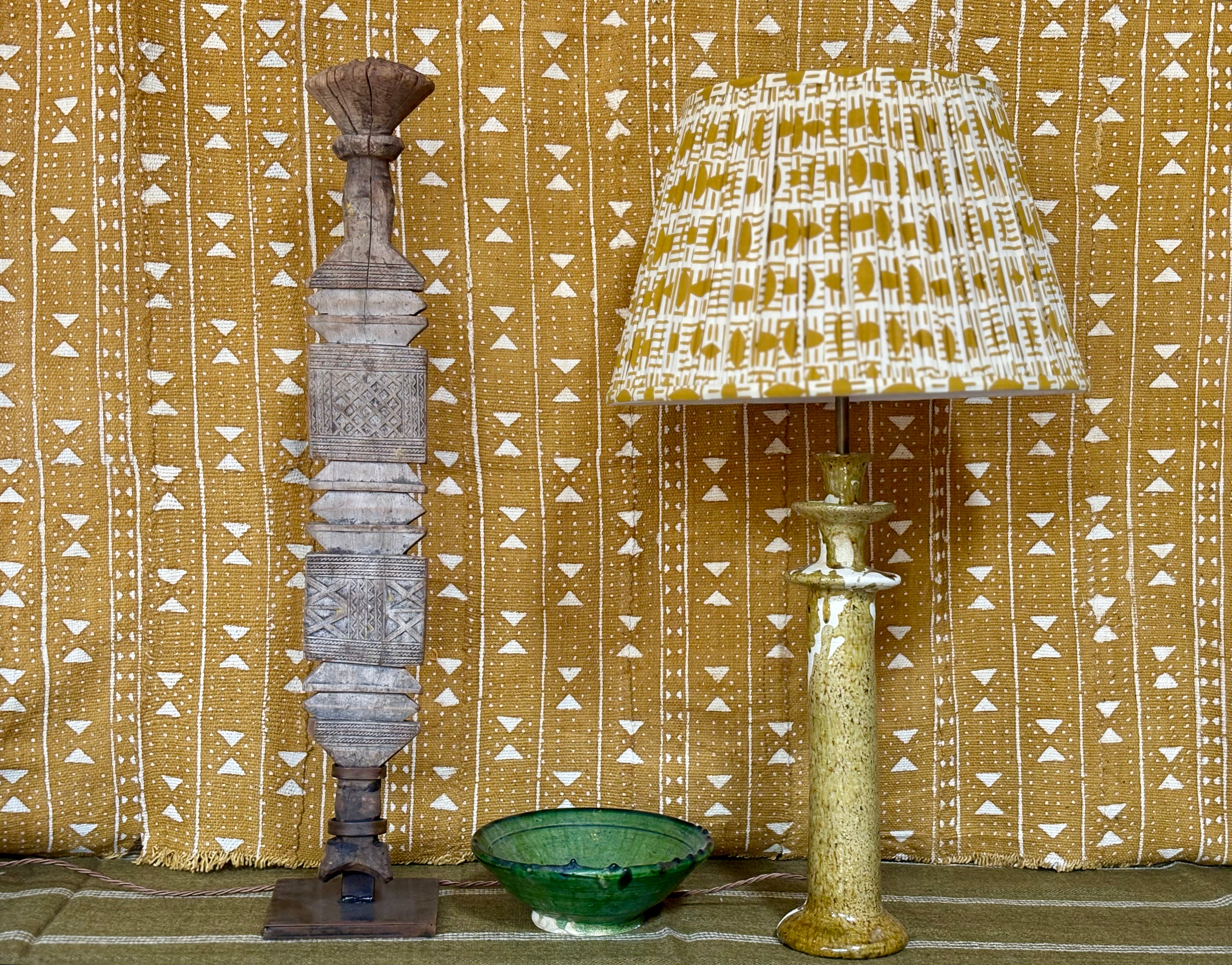 A styled collection of home decor items including a tall carved wooden pillar, a table lamp with a patterned yellow and white shade and a speckled gold base, and a small green ceramic bowl, set against a yellow and white patterned textile backdrop.
