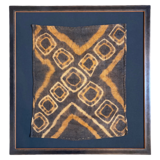 A square framed textile artwork featuring a dark charcoal background with a tan and ochre geometric diamond pattern, set within a black background mount and a dark brown wooden frame.