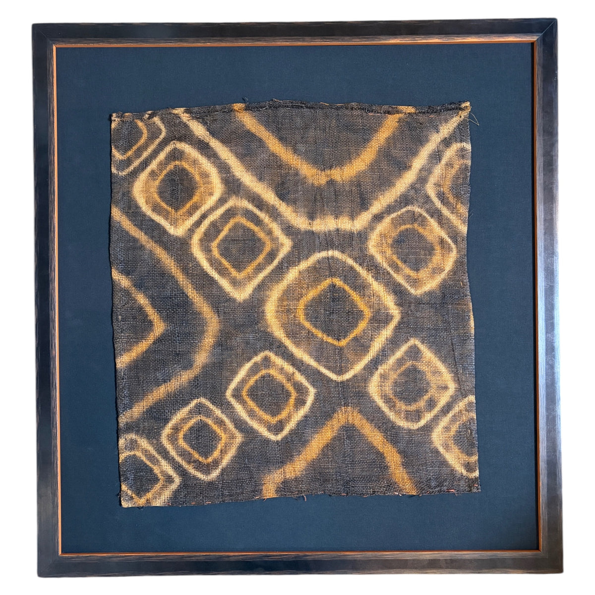 A square framed textile art piece featuring a dark charcoal background with a tan and ochre geometric diamond pattern, set in a dark wood frame with a black mat mount.