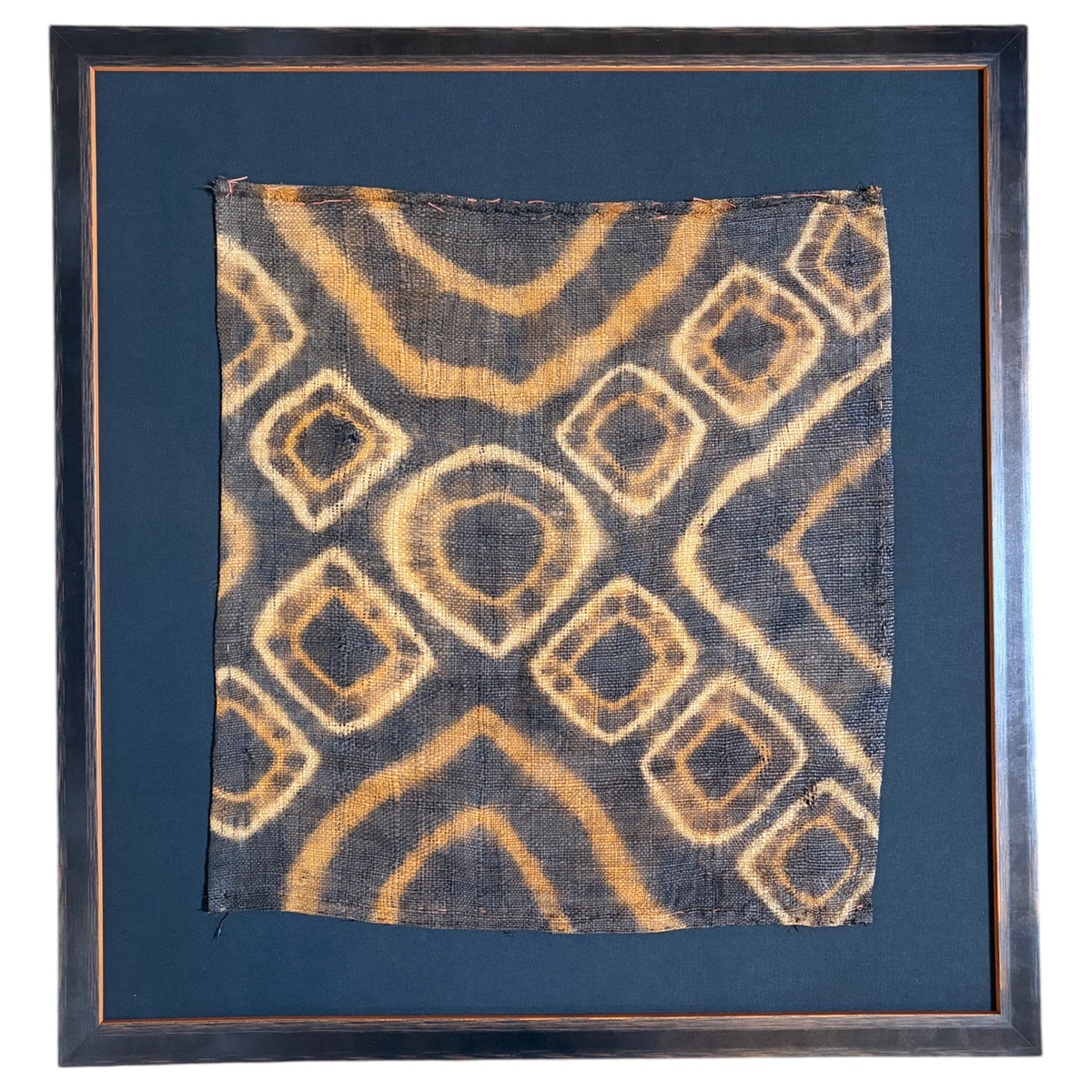 A square framed textile artwork featuring a dark charcoal fabric with a tan and beige geometric pattern, set within a dark wood frame and black mount. 