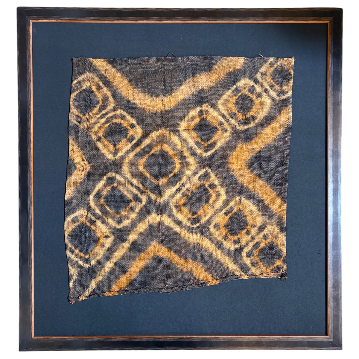 A square framed textile artwork featuring a dark charcoal fabric with a tan and ochre geometric diamond pattern, mounted on a black background with a dark wood frame.