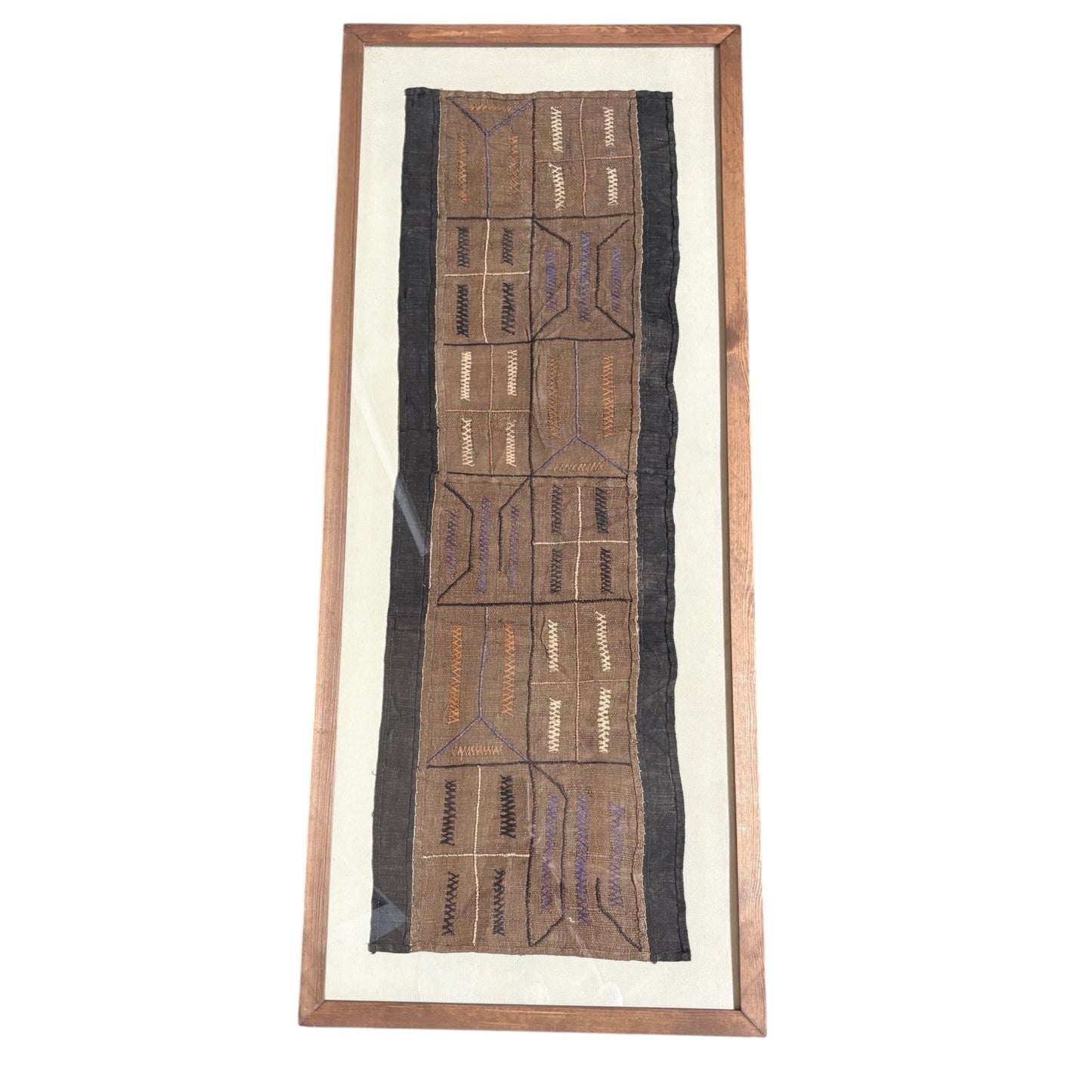 A framed African tribal textile wall hanging with geometric embroidered patterns in brown, black, and purple on a cream background, enclosed in a wooden frame.