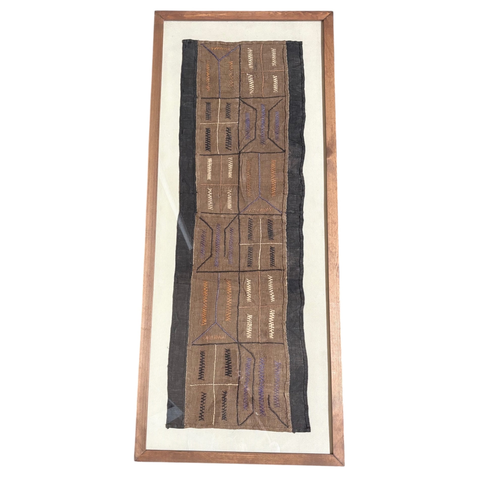 A framed African tribal textile wall hanging with geometric embroidered patterns in brown, black, and purple on a cream background, enclosed in a wooden frame.