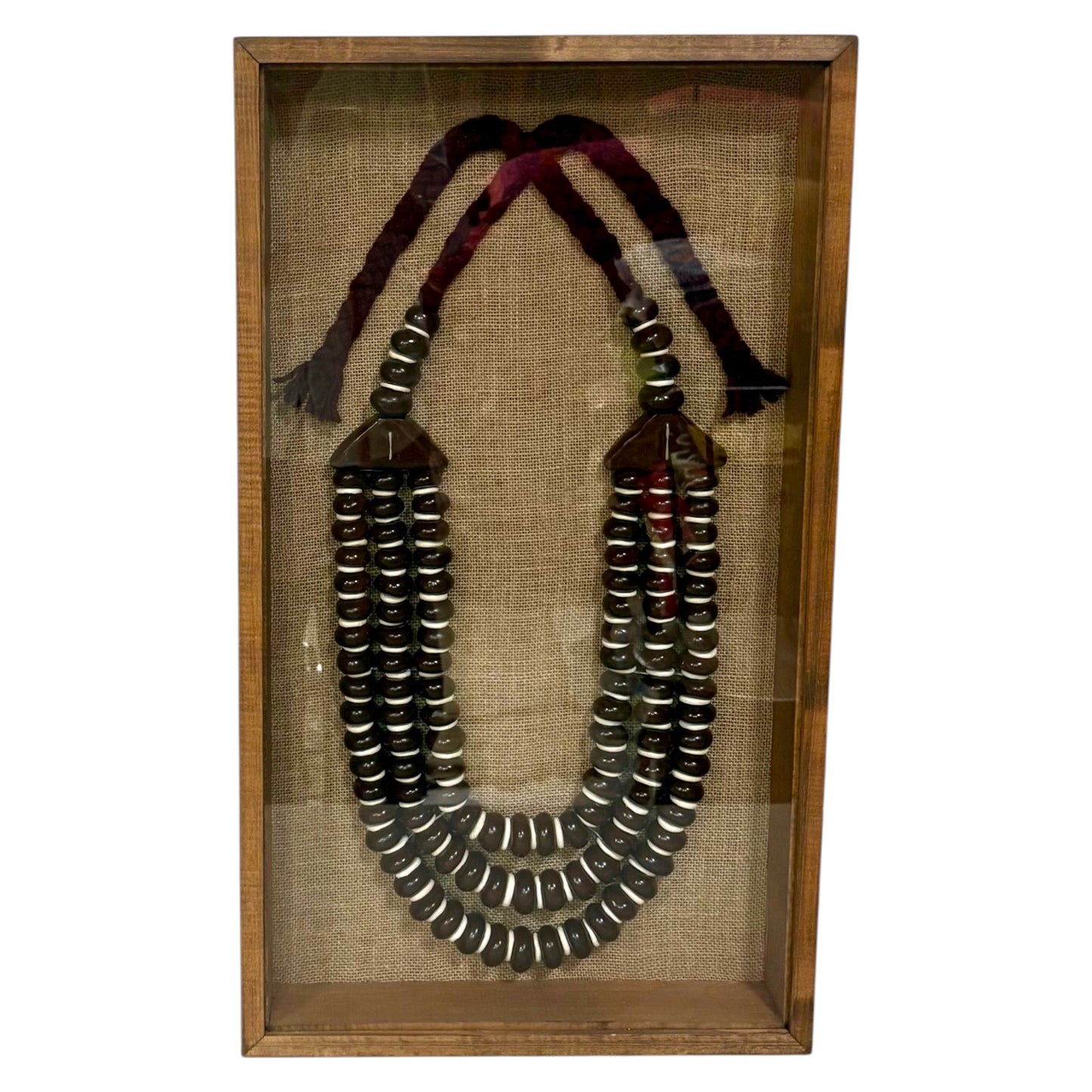 A multi-strand beaded necklace displayed inside a wooden shadow box with a Hessian background. The necklace features dark metallic beads with silver bands, dark brown wooden connector pieces, and two braided cords in dark red and black at the top.