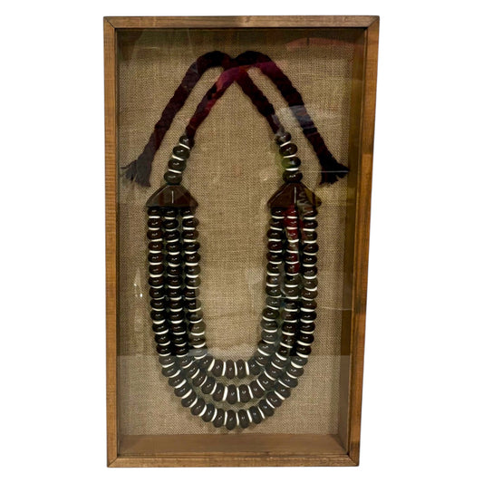 A multi-strand beaded necklace displayed inside a wooden shadow box with a Hessian background. The necklace features dark metallic beads with silver bands, dark brown wooden connector pieces, and two braided cords in dark red and black at the top.