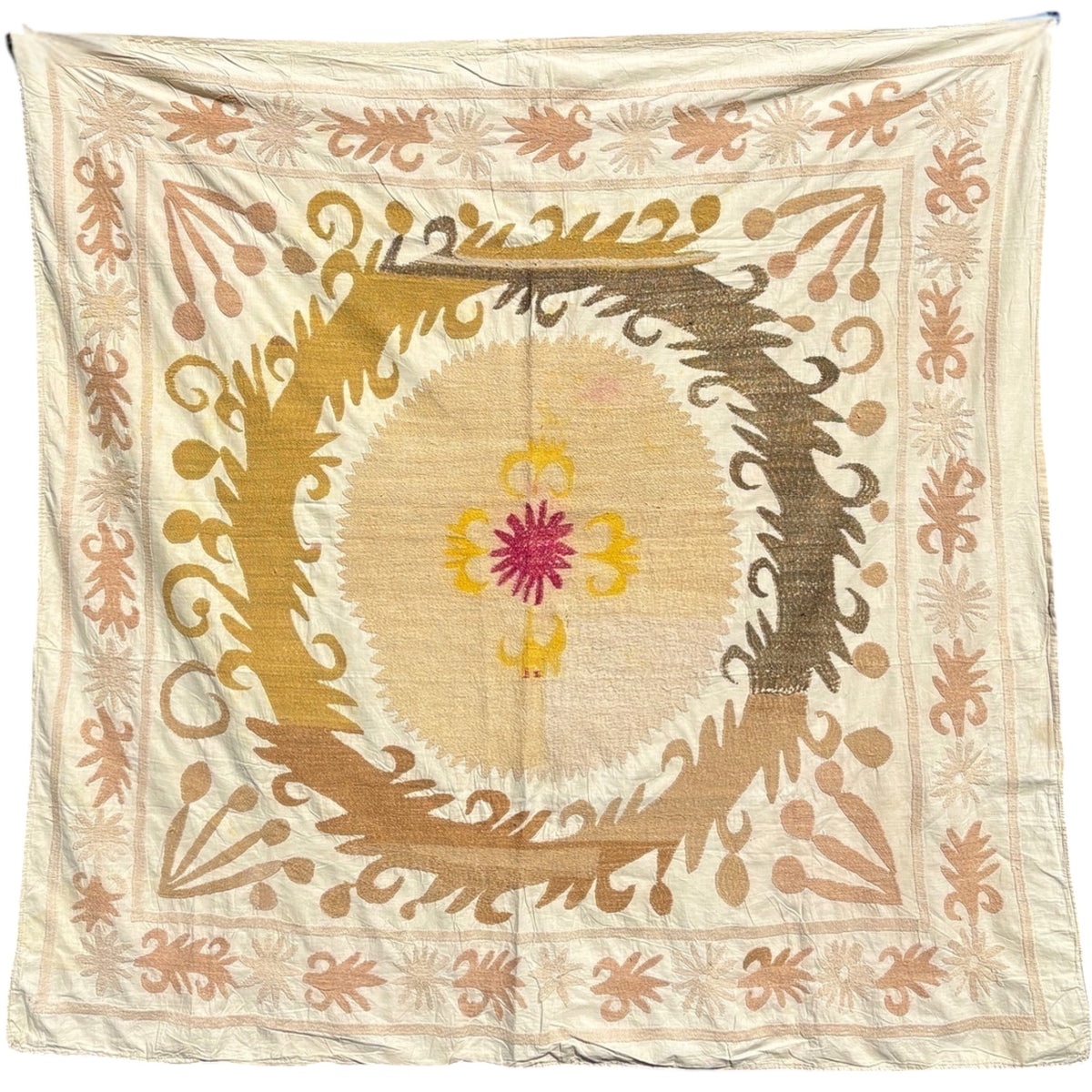 Square cream Uzbek suzani textile with a large central sunburst medallion, magenta and yellow flower, curving mustard and brown leaf scrolls, and a embroidered leaf and rosette border, hanging against a white background