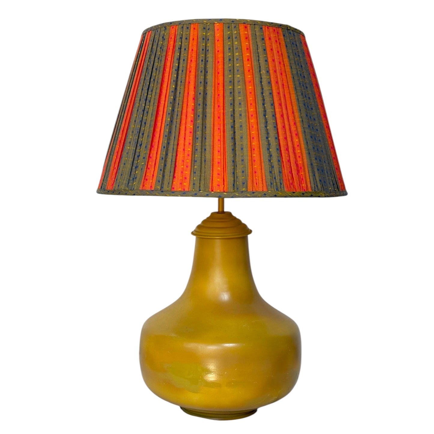 A gathered cotton fabric lampshade featuring vertical stripes of orange and green with small yellow, blue and red dotted accents, shown against a plain white background. Shown on a mustard yellow lampbase. 