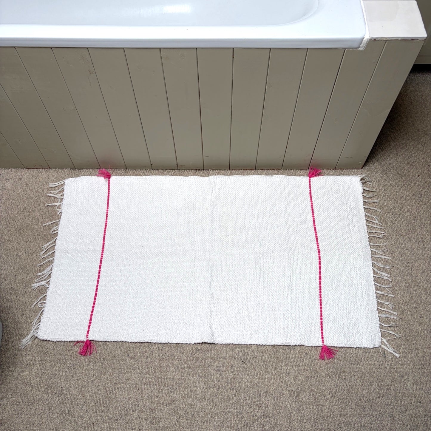 A white rectangular bath mat with fringe edges and bright pink vertical stripe accents, placed on a beige carpet in front of a bathtub.