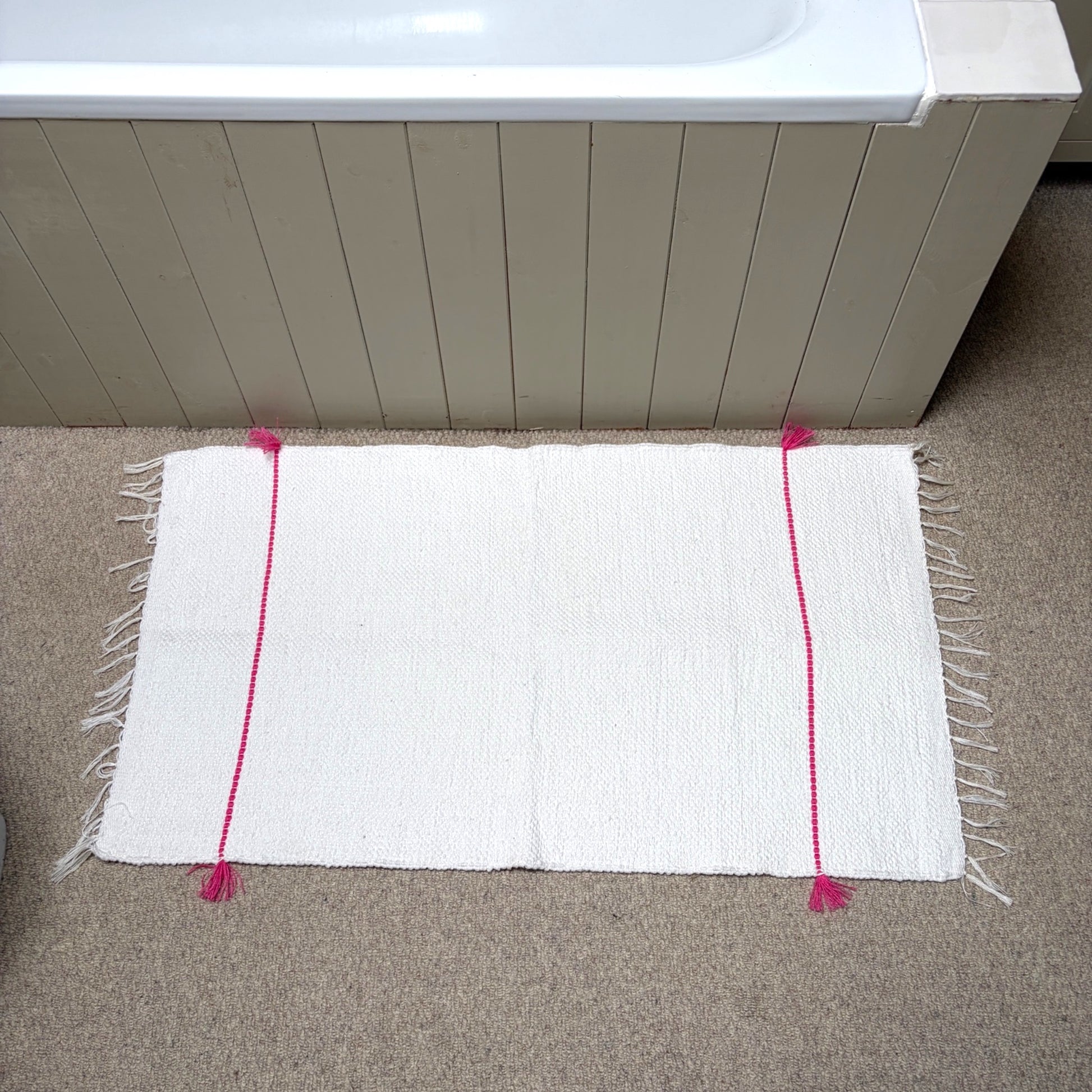 A white rectangular bath mat with fringe edges and bright pink vertical stripe accents, placed on a beige carpet in front of a bathtub.