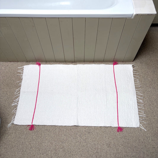 A white rectangular bath mat with fringe edges and bright pink vertical stripe accents, placed on a beige carpet in front of a bathtub.