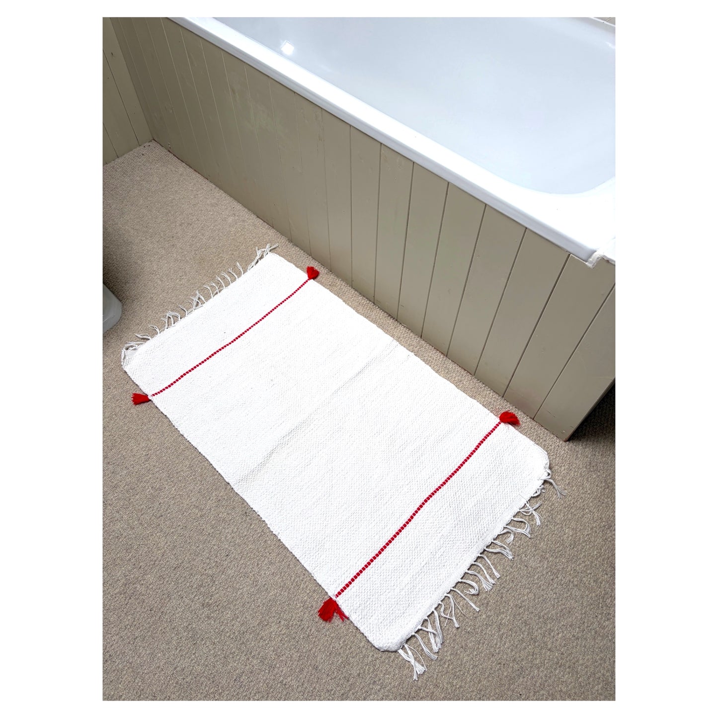 A white bath mat with red stitched lines and fringed edges, laid out on a beige carpet next to a bathtub.