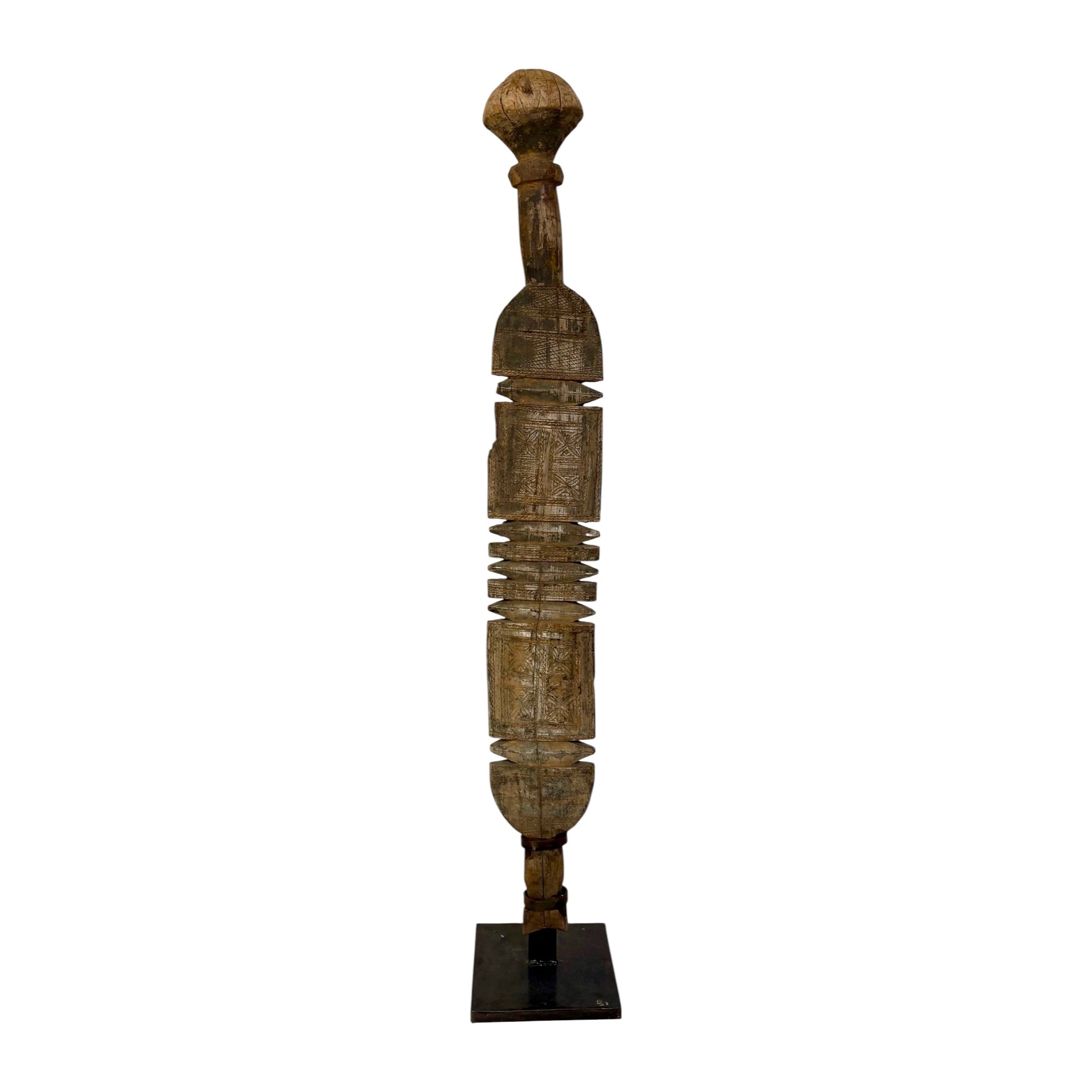 A tall, slender carved wooden sculpture with a rounded top and a dark rectangular base, photographed against a plain white background.