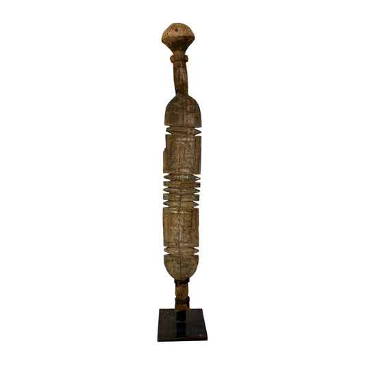 A tall, slender carved wooden sculpture with a rounded top and a dark rectangular base, photographed against a plain white background.