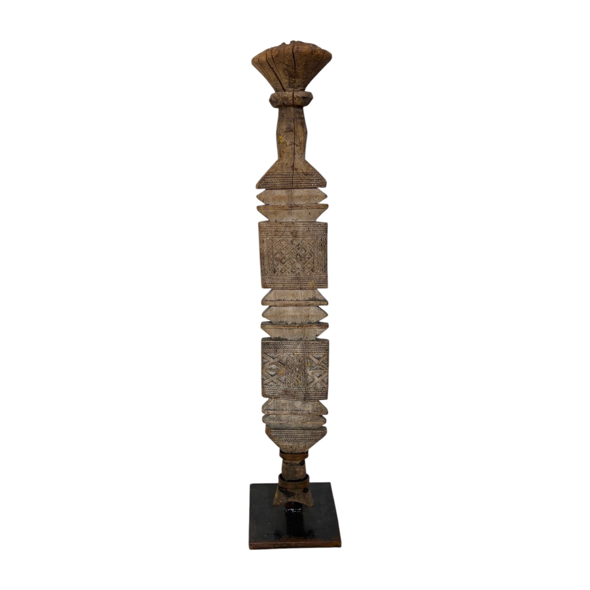 A tall, narrow carved wooden decorative pillar or finial with layered geometric and floral motifs, standing on a dark square base against a plain white background.
