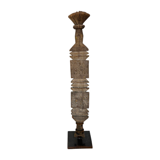 A tall, narrow carved wooden decorative pillar or finial with layered geometric and floral motifs, standing on a dark square base against a plain white background.