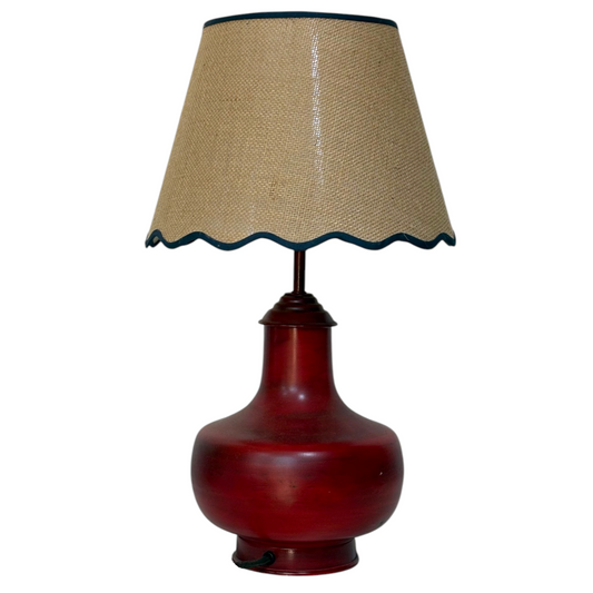 A table lamp with a glossy dark red ceramic or glass bulb-shaped base and a beige hessian lampshade with a dark blue scalloped edge, isolated on a white background.