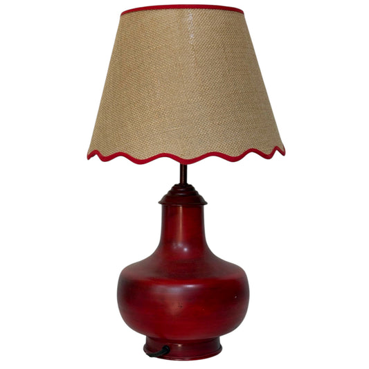A table lamp with a glossy dark red ceramic base and a beige hessian shade trimmed with a scalloped edge in red fabric.