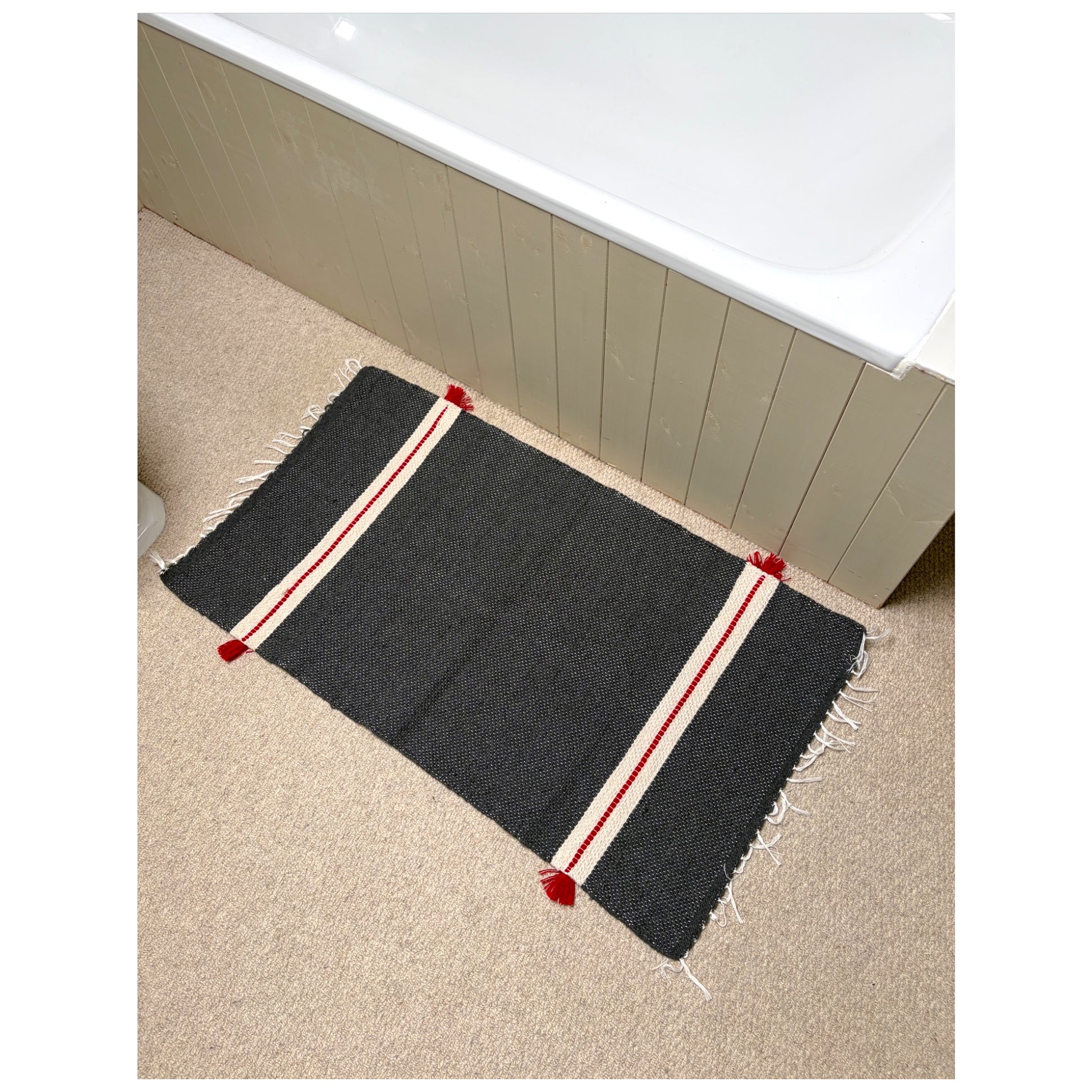 A rectangular dark gray bath mat with red and cream stripes and fringed edges, placed on a beige carpet next to a white bathtub.
