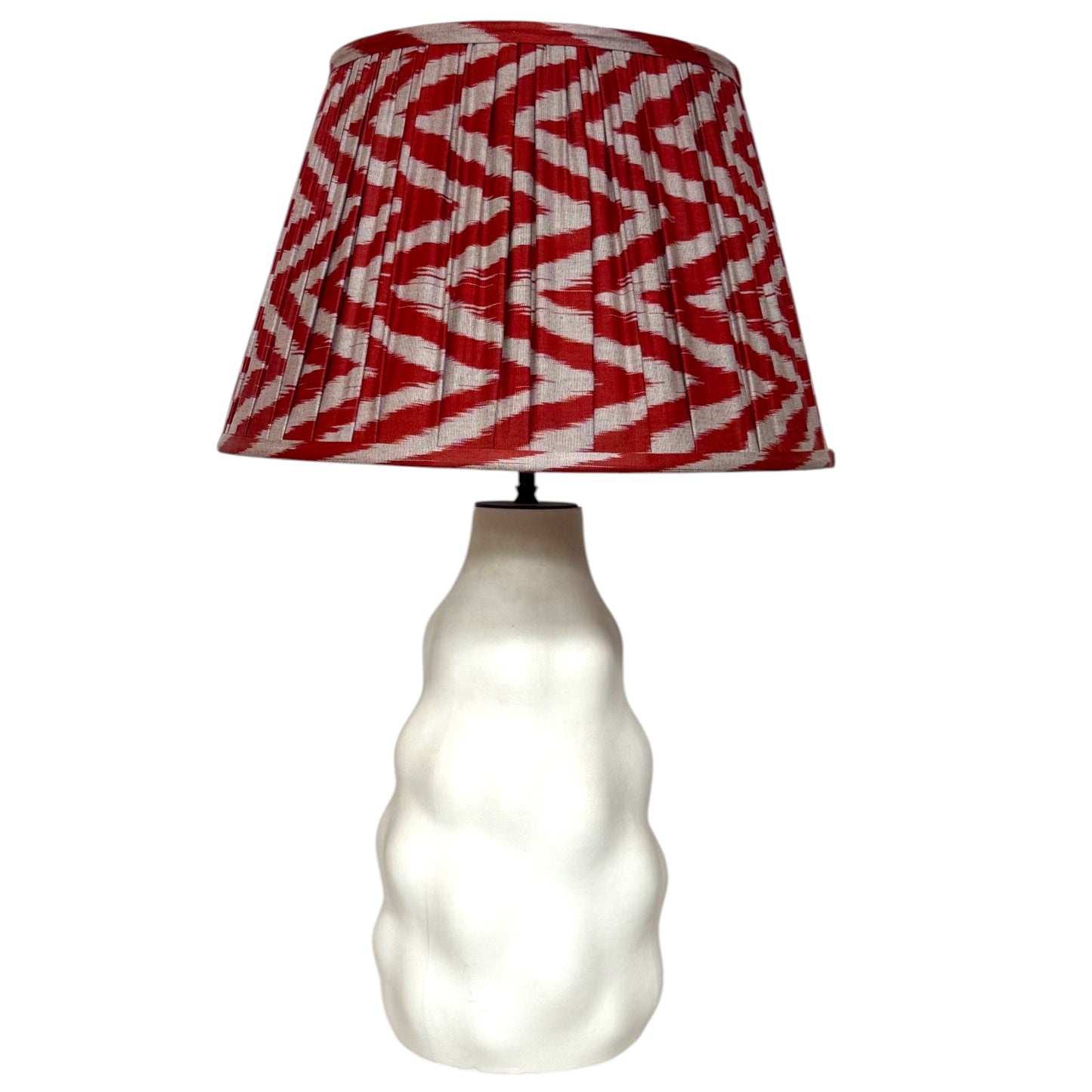 A table lamp with a smooth white ceramic base and a pleated drum shade featuring a red and white zigzag ikat pattern, set against a plain white background.