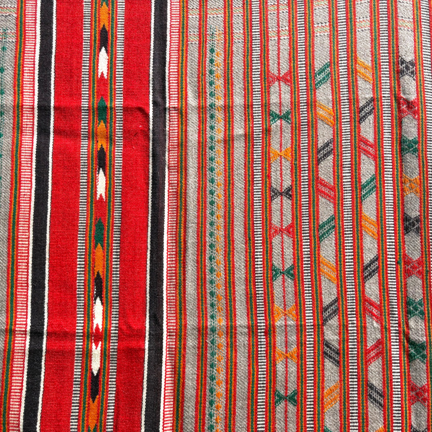 A close-up of a woven textile with vertical stripes and geometric patterns in red, black, white, orange, green, and gray.
