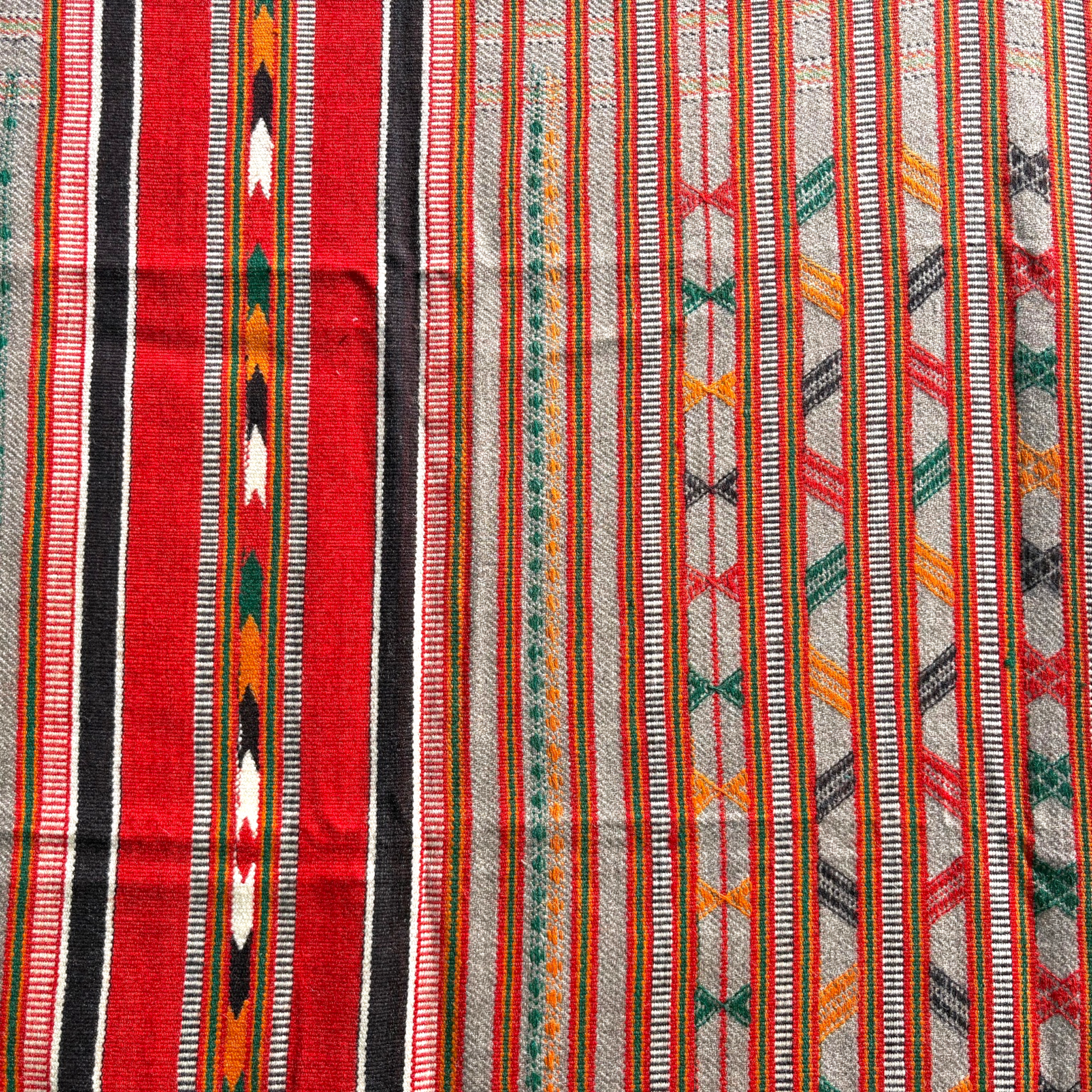A close-up of a woven textile with vertical stripes and geometric patterns in red, black, white, orange, green, and gray.