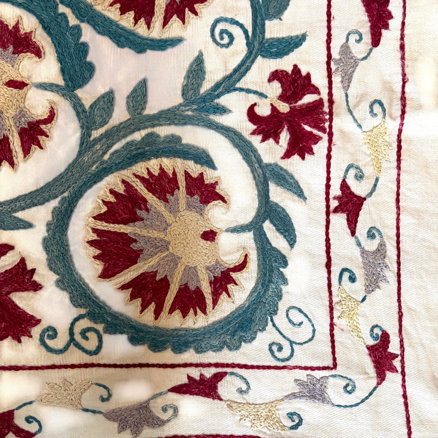 Close-up of an embroidered textile pattern featuring a central floral motif in deep red, teal blue, and beige on an off-white fabric background.