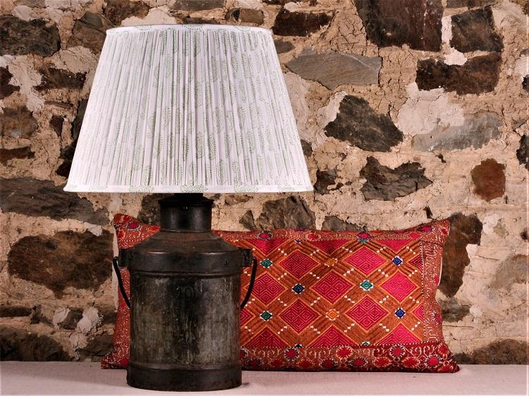 Green and White Cotton Lampshade shown on a rustic metal lamp base with e red patterned pillow in the background, against a stone wall